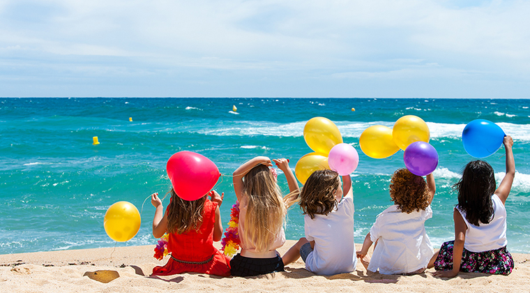 A group of children sit on the beach facing the water with their backs to us. They are holding multi-colored balloons