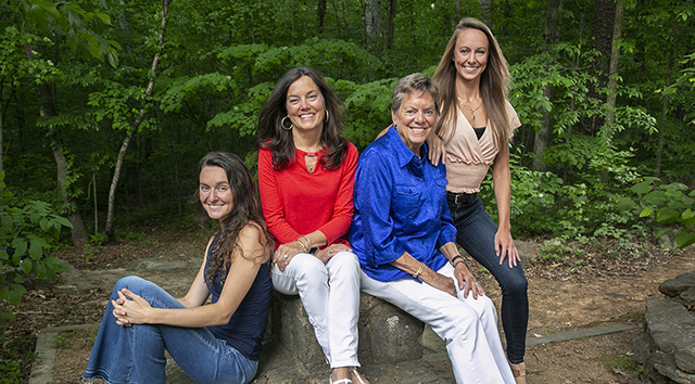 Four women pose together in a forested setting: two granddaughters, their mother, and the matriarch/grandmother. 
