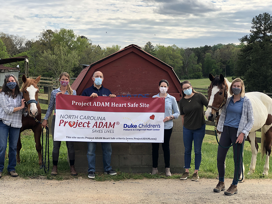 A group of people and two horses stand at an equestrian farm; they all wear masks and two hold a sign that reads "Project ADAM Heart Safe Site."