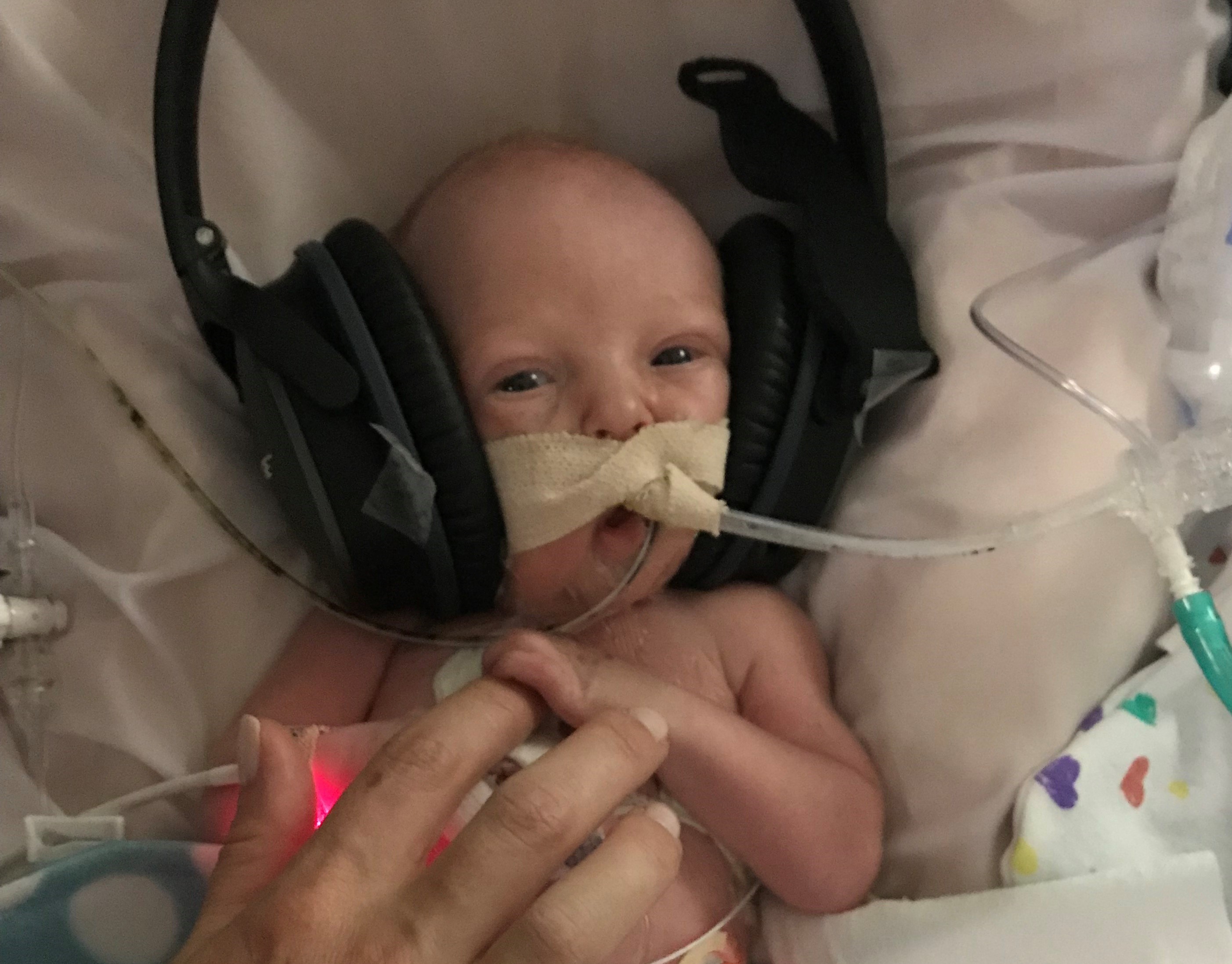 A premature baby hooked up to tubes and wires sits listening to music on giant headphones that dwarf his little head.
