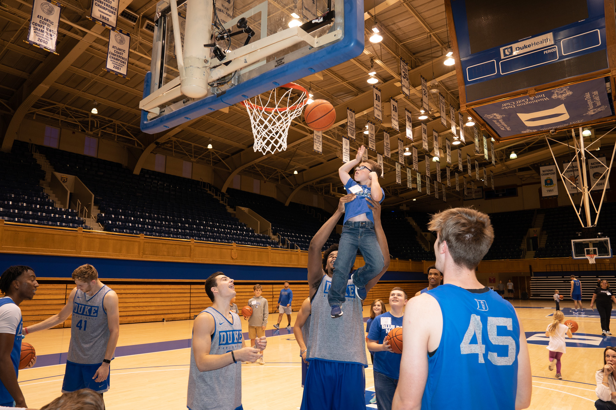 Duke Men's Basketball Players lift a young Duke Children's patient to make a basket at Cameron Indoor Stadium.
