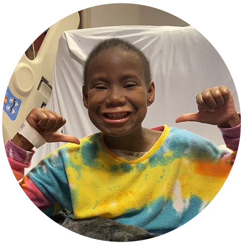 A young Black girl in a tie-dye shirt smiles and points her thumbs at herself; she is in a hospital setting. 