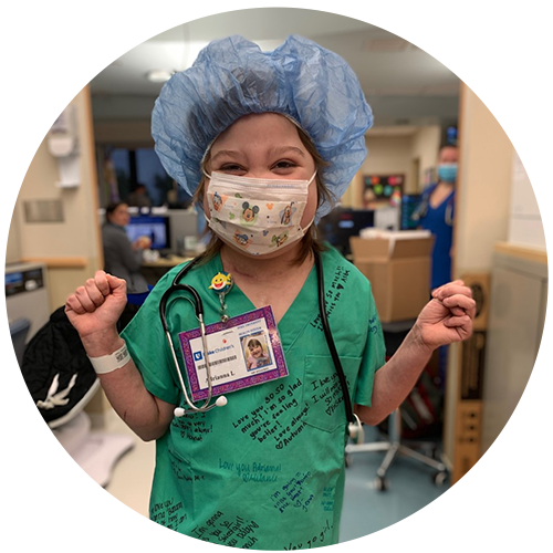A little girl in scrubs and a nurse's cap and toy stethoscope waves at the camera; she is wearing a face mask but appears to be smiling behind the mask. She is in a hospital setting. 