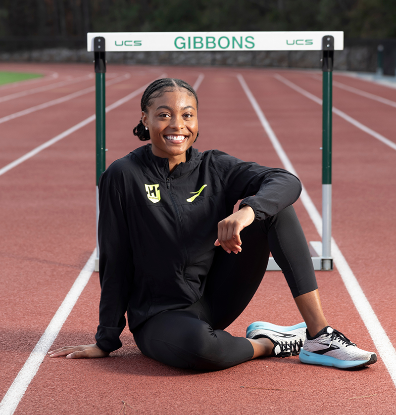 Taylor McKinnon, a young Black female athlete, poses by a hurdle on her high school's track. 