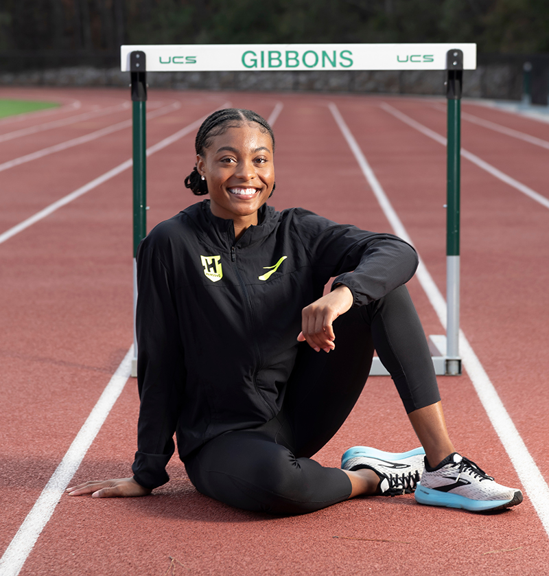 Taylor McKinnon, a young Black female athlete, poses by a hurdle on her high school's track.