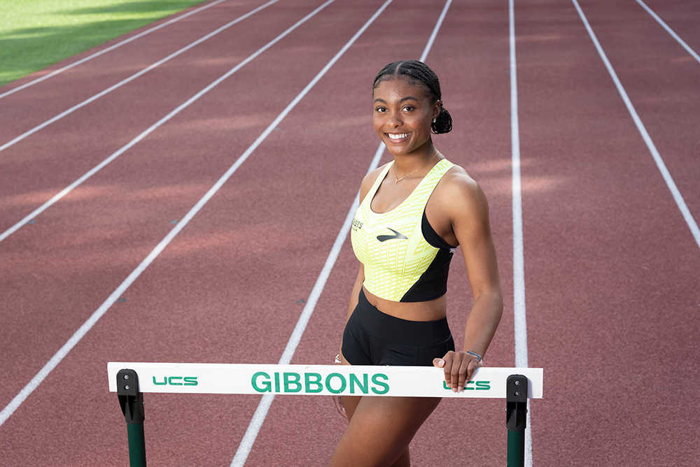 Taylor McKinnon, a young Black female athlete, poses behind a hurdle on her high school's track.