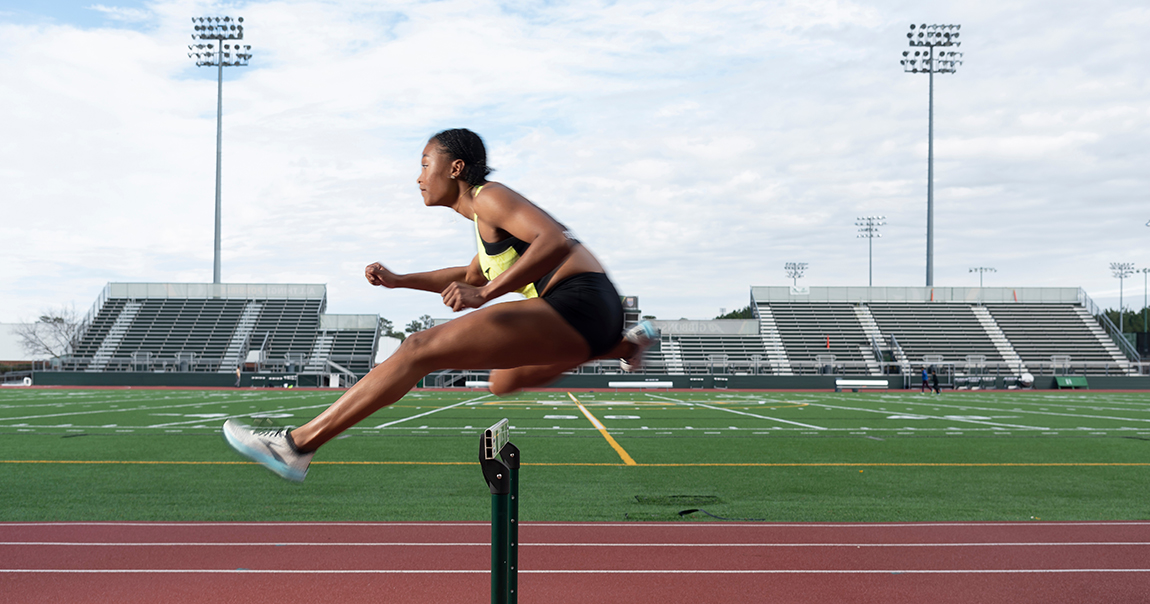 Taylor McKinnon, a young Black female athlete and high schooler, clears a hurdle at her high school's track. 