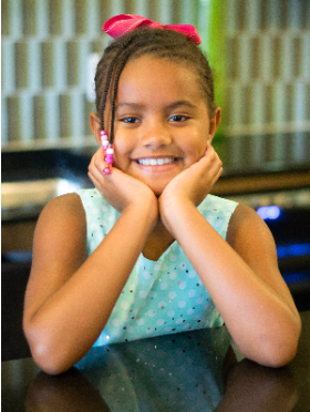 Little girl in an aqua shirt wearing a pink bow in her hair cradling her chin in her hands and a big smile