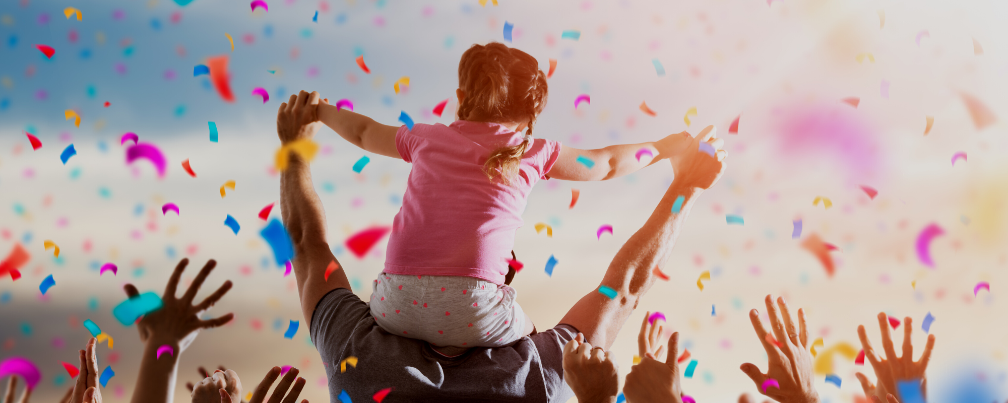 Child sitting on the shoulders of an adult with confetti falling and hands reaching upwards