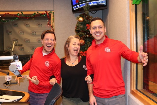 Two men in red shirts with a MIX 101.5 logo on the chest, and a woman in the middle wearing a black shirt with the same logo. They are smiling enthusiastically and standing in front of a Christmas tree. 