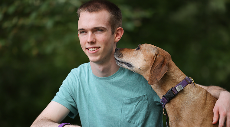A photo of Phillip Jordan, a 19-year-old white male in a teal shirt, and his dog, a tan medium-sized breed with short hair and floppy ears.