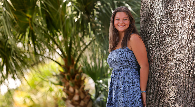 Sarah Drury, a 15-year-old girl with tan skin and brown hiar and brown eyes, stands next to a tree and smiles at the camera. She wears a pale blue strappy dress with blue polka dots.