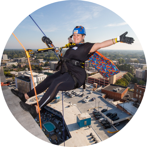 A woman stands at the edge of 21c Museum Hotel in downtown Durham; she is about to rappel down the building. She wears black athletic garb and a helmet, her arms are outstretched and she is smiling. 