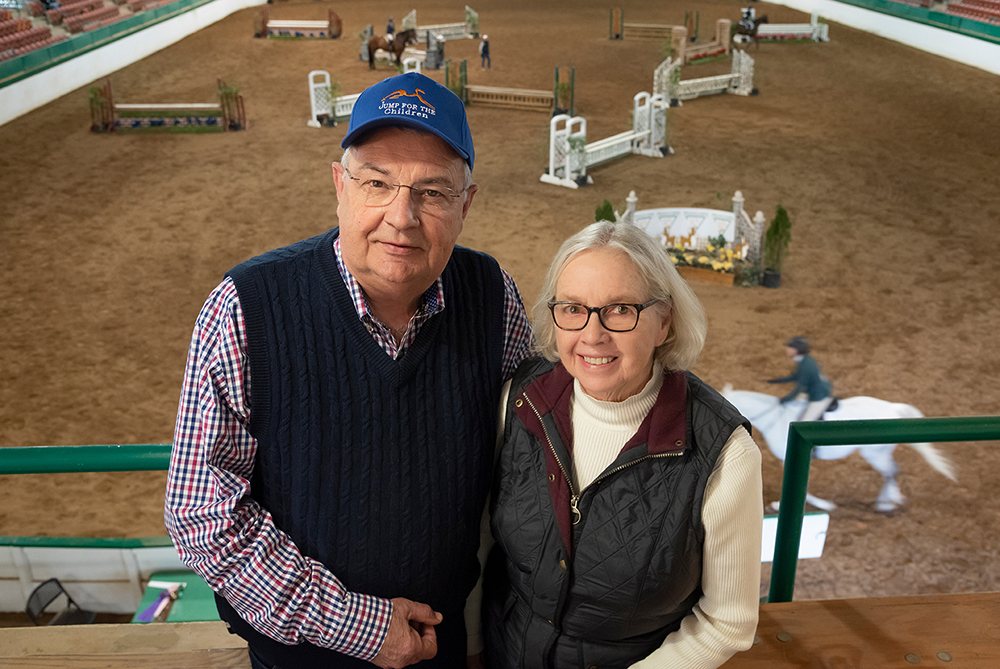 Glenn and Joan Petty stand in front of the riding arena at the James B. Hunt Horse Complex at the State Fairgrounds in Raleigh. Joan and Glenn are white individuals in their 70s. Both wear glasses and casual riding gear. Glenn wears a blue had that says "Jump for the Children." 