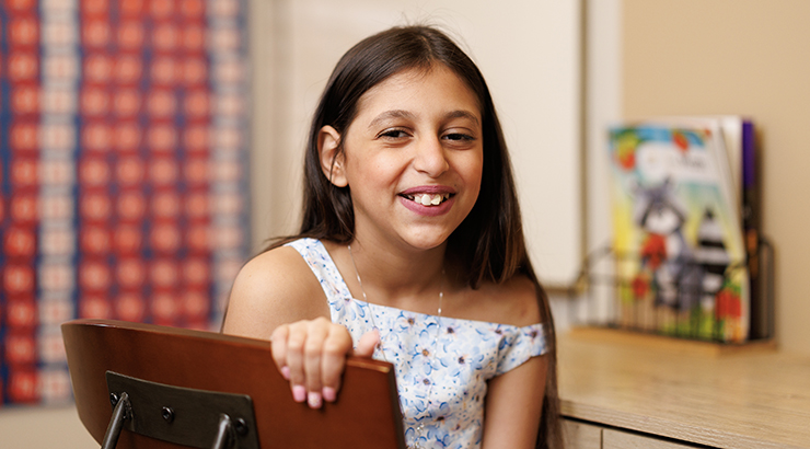Ela Allam smiles at the camera; she is a 9 or 10 year old girl with brown hair and eyes and olive skin. She is sitting in her bedroom at a desk. 