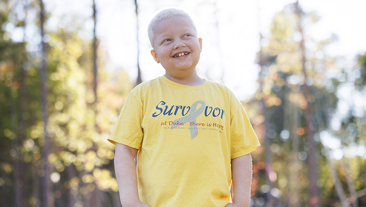 Randall Saladin, an 8-year-old boy with bleach blonde hair, wears a yellow shirt that says "survivor" on it while looking off into the distance and smiling.