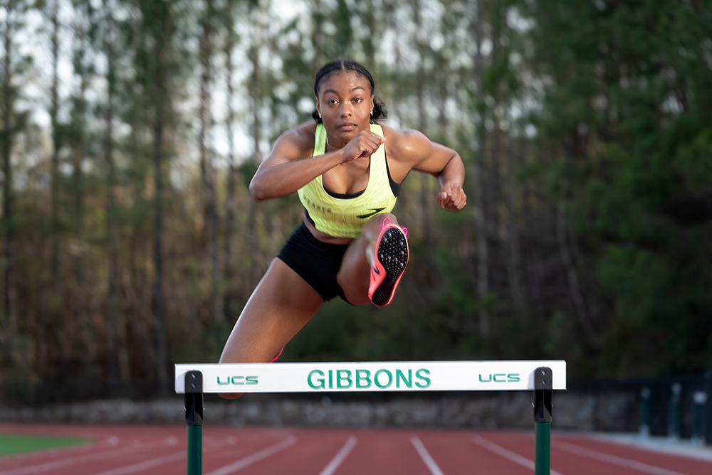 Taylor McKinnon, a young Black female athlete, clears a hurdle on her high school's track.