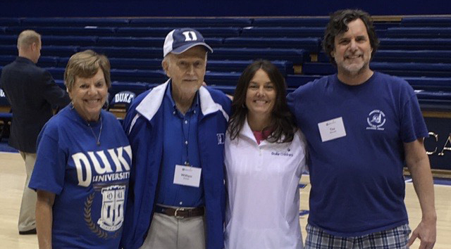 Four people stand in Cameron Indoor Stadium, Duke's Basketball Stadium: an older woman, an older man, a middle-aged woman, and a middle-aged man. All wear blue and white Duke athletics fan gear. The older man wears a baseball cap.