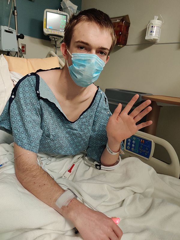 Phillip Jordan in the ICU; he wears a hospital gown and a face mask and waves at the camera.