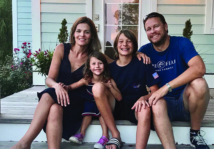 A family of four sits together on their doorstep -- a mom, a dad, a big brother, and a little sister. All are dressed in shades of navy and all are smiling. 