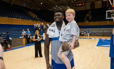 Zion Williamson, Duke Men's Basketball Player, holding a child on a basketball court.