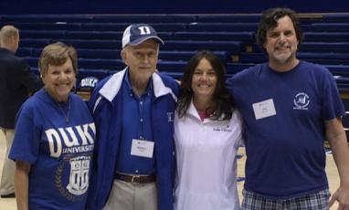 Four people stand in Cameron Indoor Stadium, Duke's Basketball Stadium: an older woman, an older man, a middle-aged woman, and a middle-aged man. All wear blue and white Duke athletics fan gear. The older man wears a baseball cap.