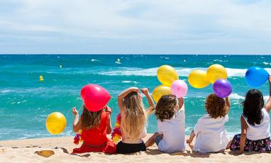 A group of children sit on the beach facing the water with their backs to us. They are holding multi-colored balloons