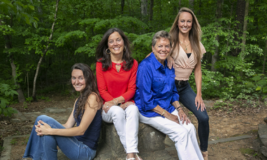 Four women pose together in a forested setting: two granddaughters, their mother, and the matriarch/grandmother.