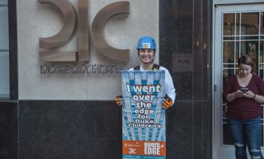 A woman stands in front of 21c Museum Hotel wearing a helmet and holding a sign that reads "I went Over the Edge for Duke Children's!"