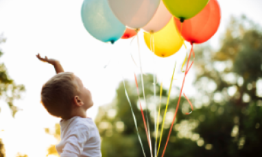Little boy reaching towards colorful balloons with sun shining through the trees behind him