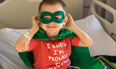 Little boy sitting in a hospital bed wearing a green cape and green mask like a super hero