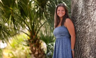 Sarah Drury, a 15-year-old girl with tan skin and brown hiar and brown eyes, stands next to a tree and smiles at the camera. She wears a pale blue strappy dress with blue polka dots.