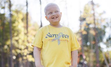 Randall Saladin, an 8-year-old boy with bleach blonde hair, wears a yellow shirt that says "survivor" on it while looking off into the distance and smiling.